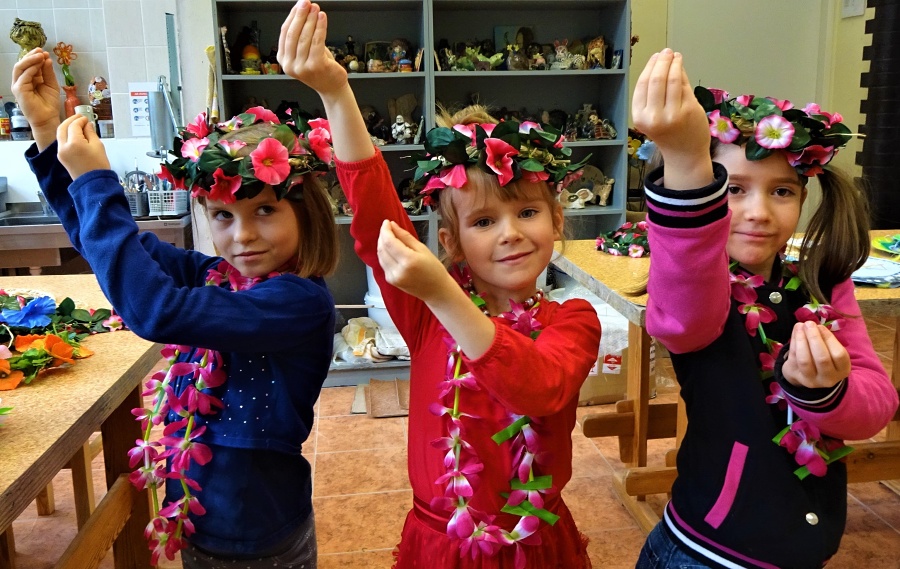 Trzy dziewczynki stoją obok siebie z rękami uniesionymi do góry tworząc z palców dłoni pąk kwiatowy. Mają na głowach wianki , a na szyi girlandy z różowych kwiatów.