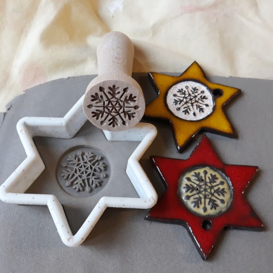 Na plastrze gliny leży foremka do ciastek, pieczątka i dwie ceramiczne choinkowe ozdoby o kształcie gwiazdki. 