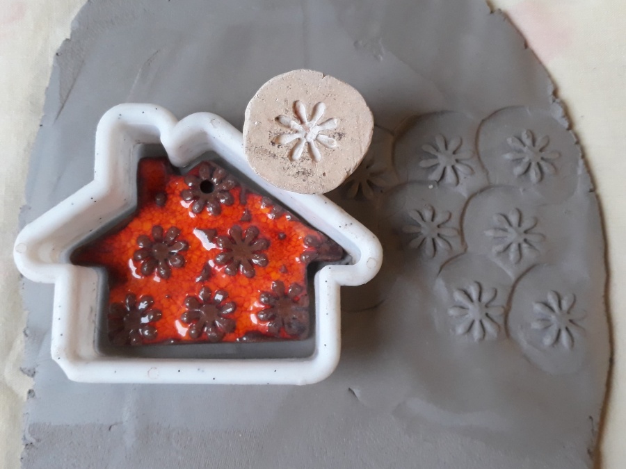 Na plastrze gliny leży ceramiczna ozdoba choinkowa o kształcie domku i pieczątka. 