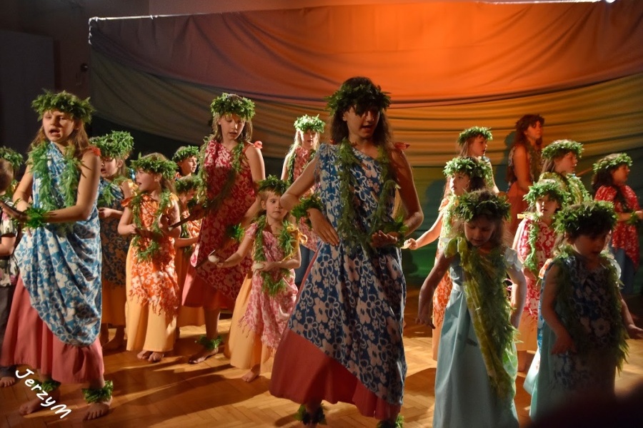 Grupa dziewcząt ubranych w kolorowe parea w białe kwiaty, w zielonych wiankach na głowach, z kijami w dłoni bądź bez, tańczy na scenie i śpiewa.
