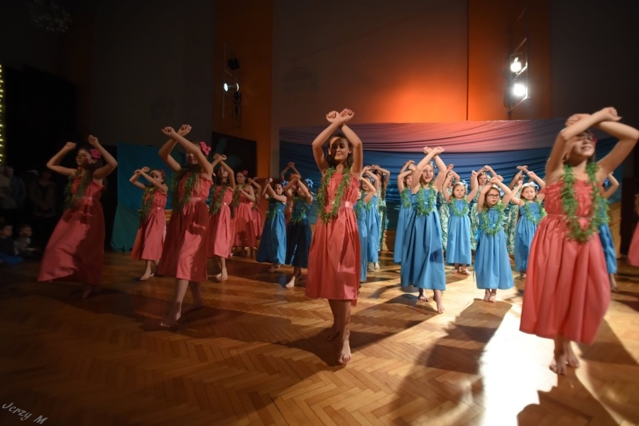 Grupa dziewcząt w różowych i turkusowych sukniach z kwiatami w tych samych kolorach tańczy na scenie trzymając ręce skrzyżowane przed sobą.