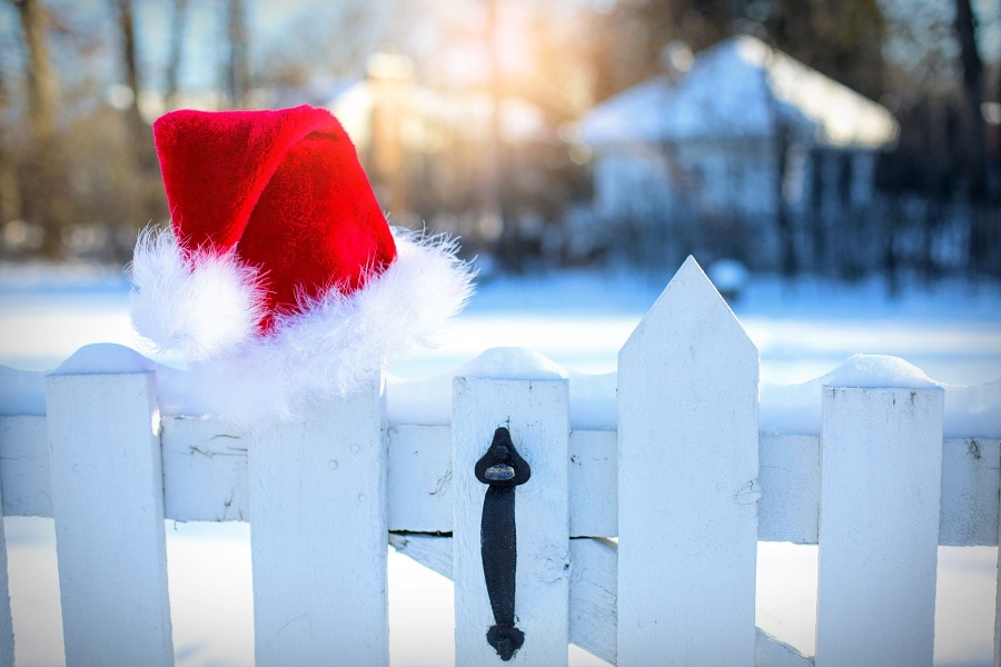 Zdjęcie przedstawia czapkę św. Mikołaja zaczepioną na białej furtce wejściowej, prowadzącej do domu w ośnieżonej scenerii.