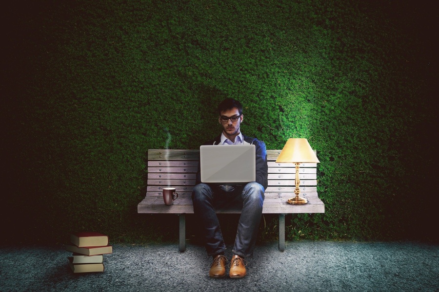 Zdjęcie przedstawia mężczyznę siedzącego na ławce, trzymającego na kolanach laptop.