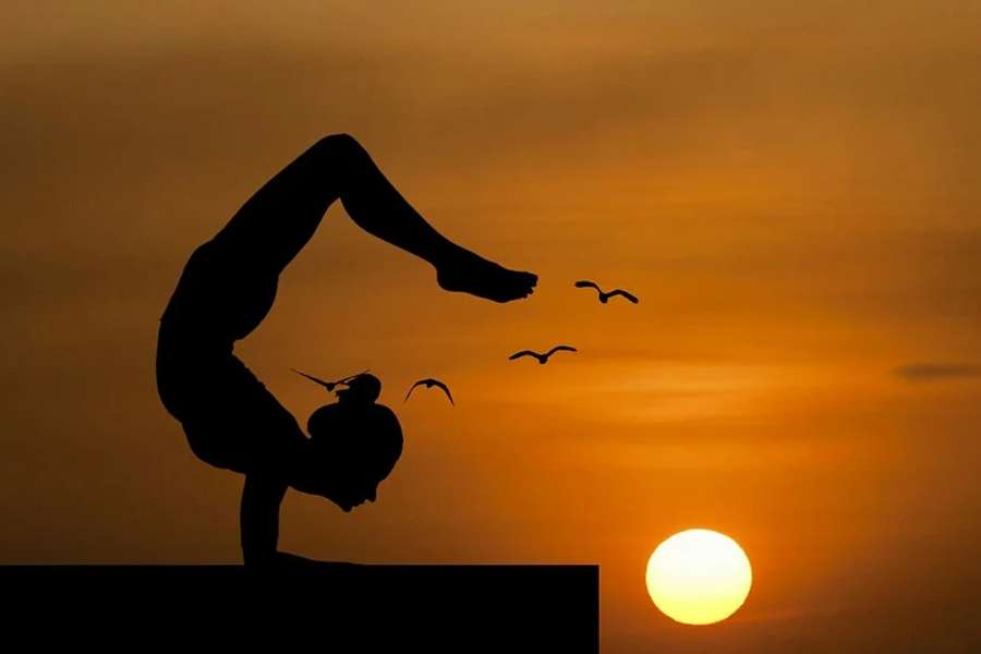 Zdjęcie przedstawia postać kobiecą ćwiczącą na tle zachodzącego słońca wśród latających ptaków.