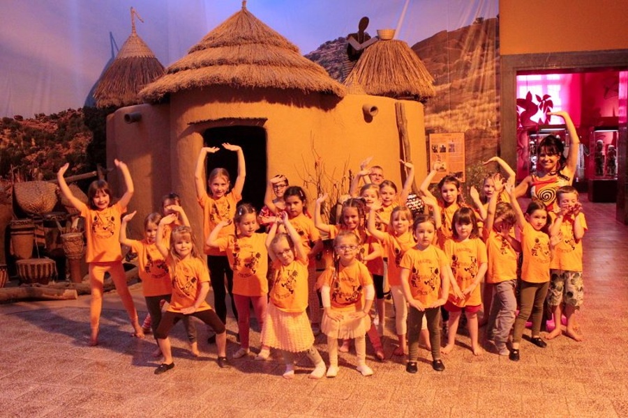 Grupa dzieci w pomarańczowych bluzkach oraz kobieta stoją w tanecznej pozie z rękami u góry przed chatami afrykańskimi.