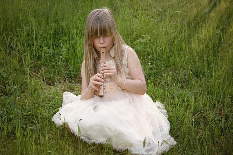 Na zdjęciu widać dziewczynkę w białej sukience,która siedzi na trawie i gra na flecie prostym.