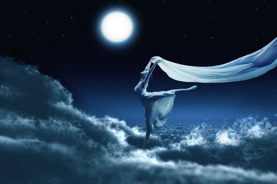 Zdjęcie przedstawia baletnicę z białą woalką stojącą w chmurach przy blasku pełni księżyca