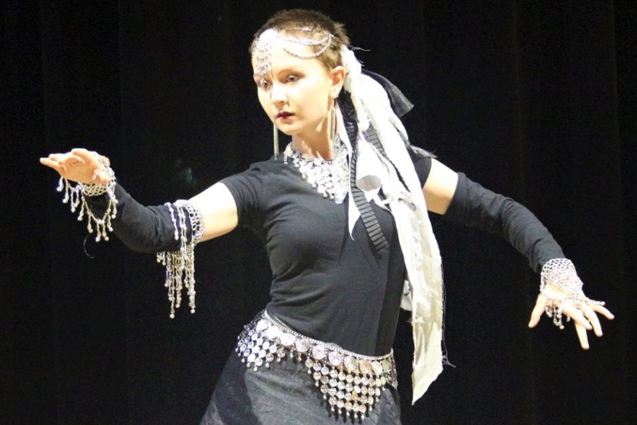 Kobieta w czarnej sukience ze srebrnym pasem i srebrnymi ozdobami na włosach i rękach stoi w tanecznej pozie na scenie.