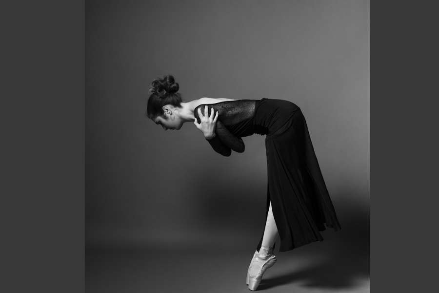 Zdjęcie przedstawia tancerkę stojącą na pointach. Sylwetka pochylona w przód, ręce splecione obejmujące ramiona.