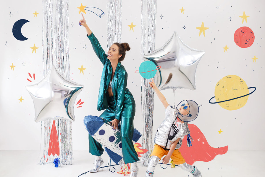 kobieta w błyszczącym zielono-turkusowym stroju i dziewczynka w srebrzystym sięgają do dekoracji z gwiazd- baloników
