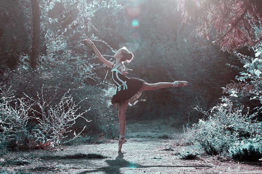 Zdjęcie przedstawia tancerkę stojącą wśród drzew i krzewów w pozie arabesque na pointach.