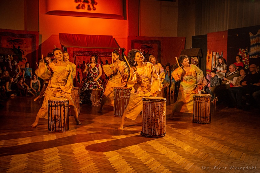 Kobiety ubrane w barwne suknie, trzymające drewniane pałki w rękach w tanecznej pozie. Przed każdą stoją bębny afrykańskie.