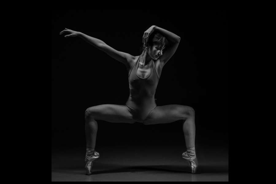 Zdjęcie przedstawia tancerkę na czarnym tle w pozycji  grand plie na pointach z prawą ręką ułożoną na głowie, lewą wyciągniętą w przód.