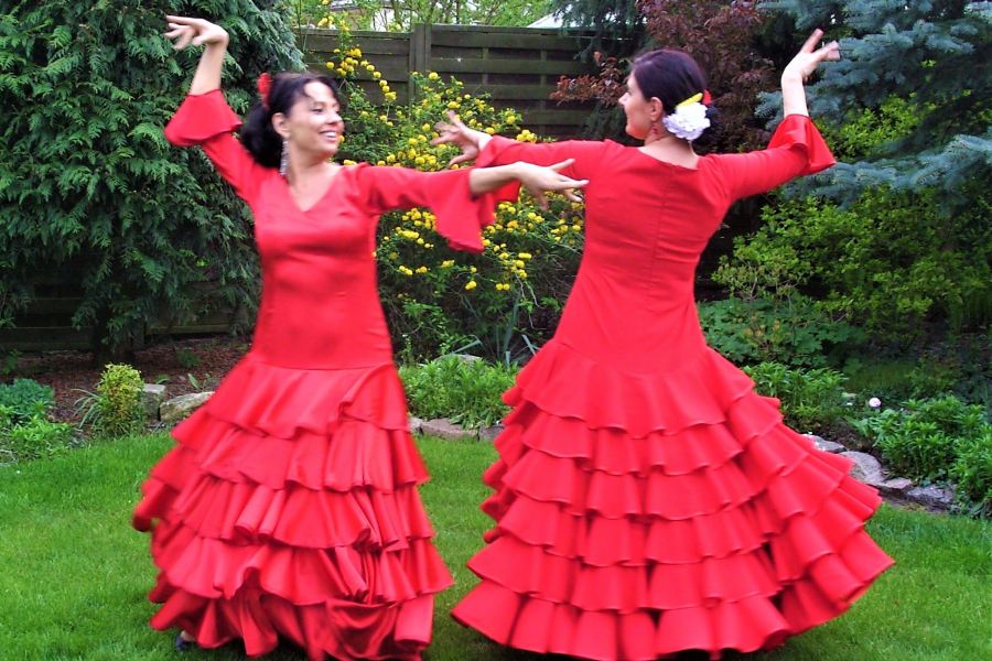 Dwie kobiety w czerwonych sukniach z falbanami na trawie w ogrodzie. Patrzą na siebie. Stoją w pozie tanecznej hiszpańskiego flameco