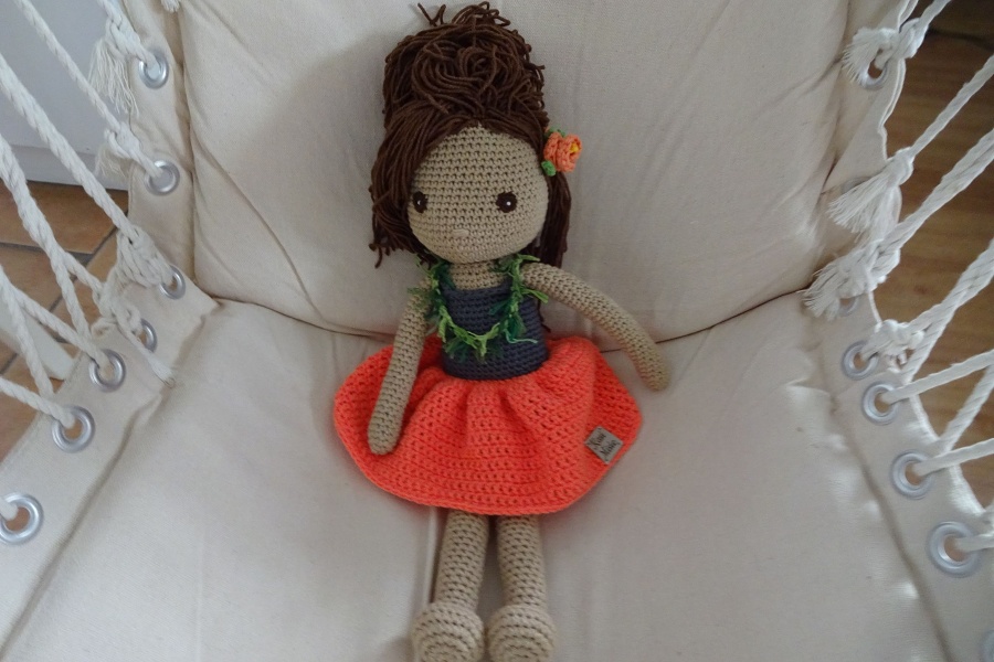 Szydełkowa lalka w pomarańczowej spódnicy, z hawajską girlandą na szyi i kwiatem we włosach siedzi na płóciennej huśtawce.