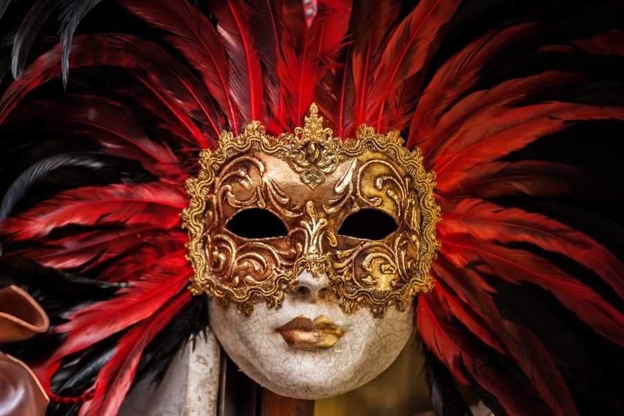 Maska wenecka o złotej i wzorzystej górnej połowie i białej dolnej połowie, złote usta, wokół maski wystające czerwone pióra, czarne tło