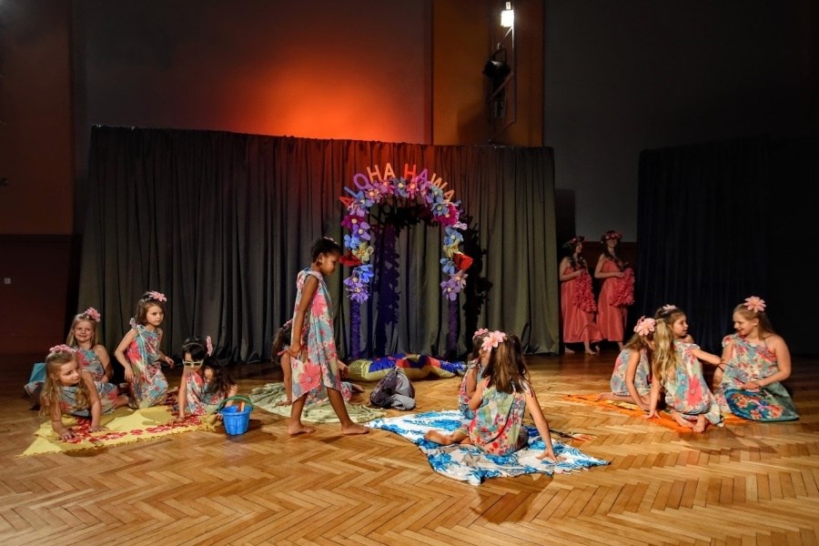 Grupa dzieci ubrana w kolorowe parea siedzi i stoi na scenie. W tle dekoracja z napisem Aloha Hawaje.