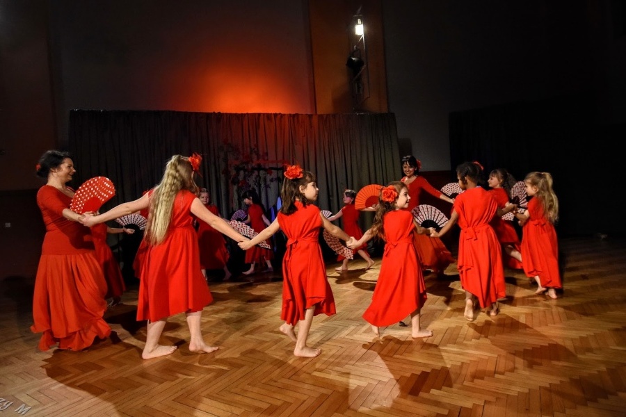Grupa dziewcząt ubranych w czerwone suknie tańczy w kole trzymając wachlarze w kropki.