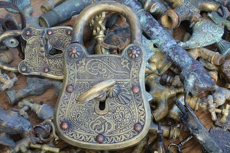 Pięknie rzeźbiona stara szarometalowa kłódka z włożonym kluczem na tle rozrzuconych kawałków metalowych innych kluczy, łańcuszków i kluczyków
