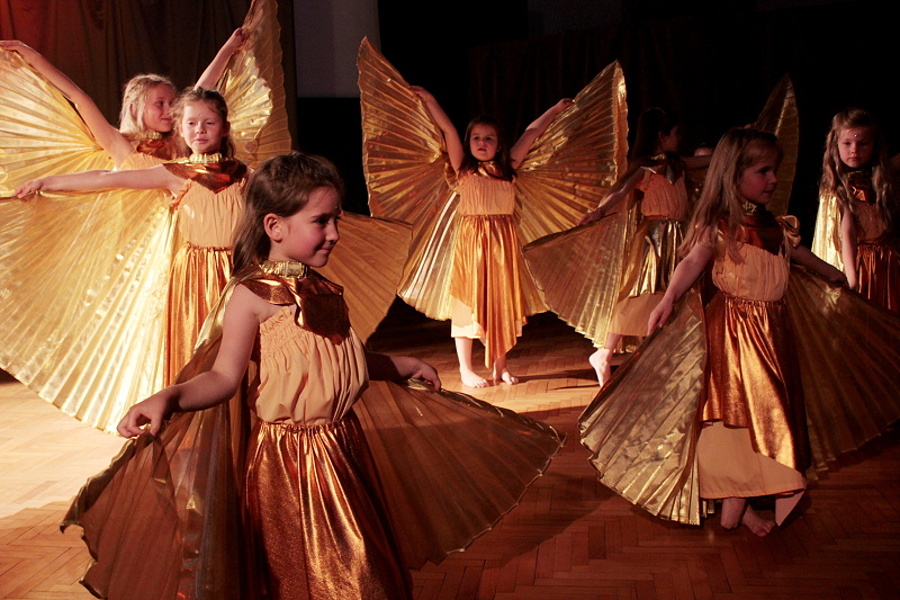 Grupa dziewcząt ubranych w złote kostiumy ze złotymi skrzydłami stoi na scenie w tanecznej pozie.