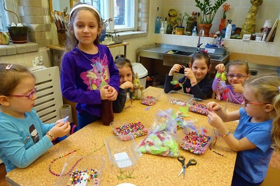 Grupa dziewcząt kolorowo ubranych siedzi przy stole i nawleka kolorowe koraliki i piórka tworząc biżuterię.