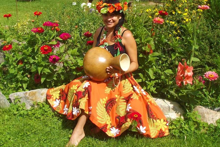 Kobieta z pomarańczowym wiankiem na głowie i w pomarańczowej spódnicy w kwiaty siedzi na trawie trzymając tykwę w dłoniach.