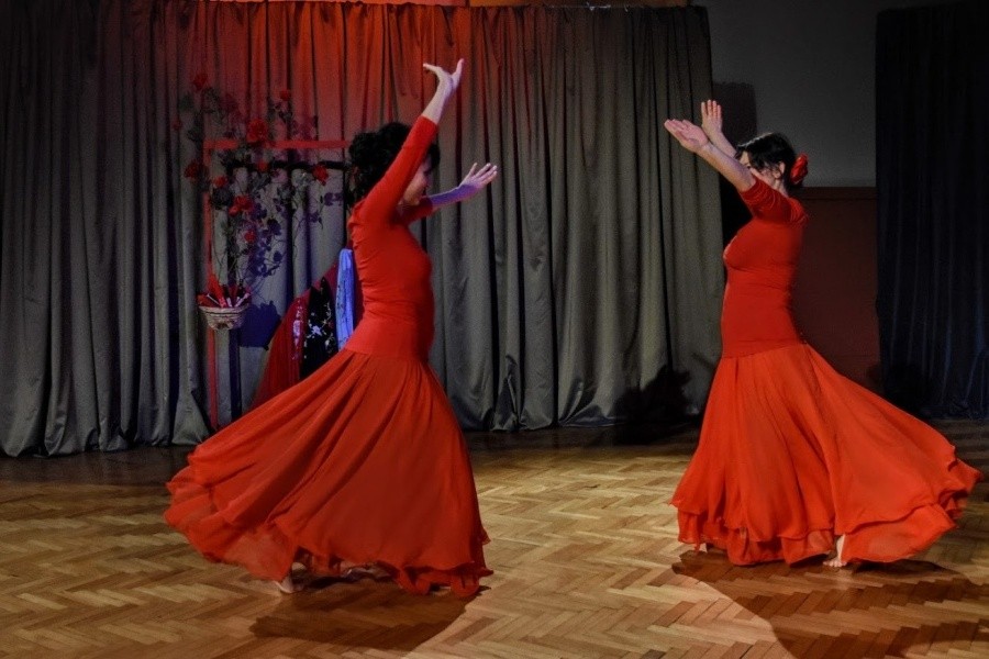 Dwie kobiety ubrane w czerwone suknie stoją w tanecznej pozie z jedną ręką u góry, przodem do siebie.