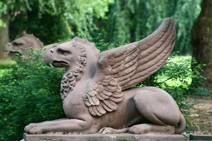 Szary kamienny posąg gryfa z lekko uniesionymi skrzydłami widziany z profilu na tle zielonego parku oraz rozmazanego posągu drugiego gryfa