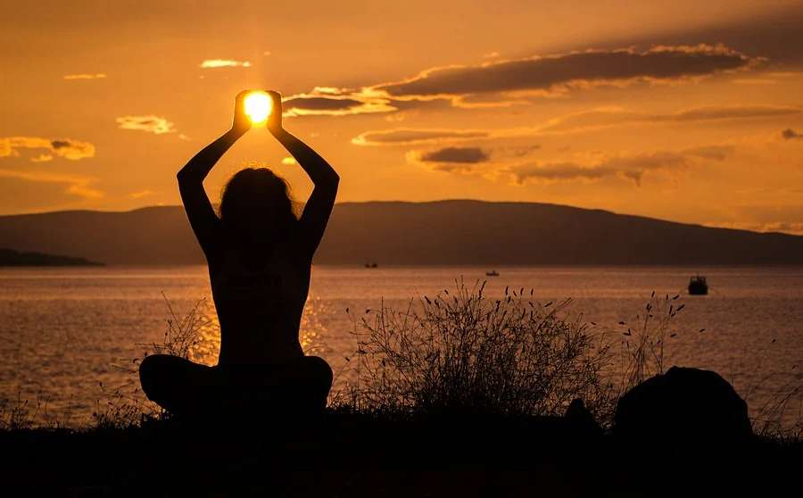 Zdjęcie przedstawia zachód słońca i postać kobiety  w pozycji  siedzącej z nogami ułożonymi po  turecku,  ręce  znajdują  się  w górze nad głową - iluzja trzymania słońca w dłoniach