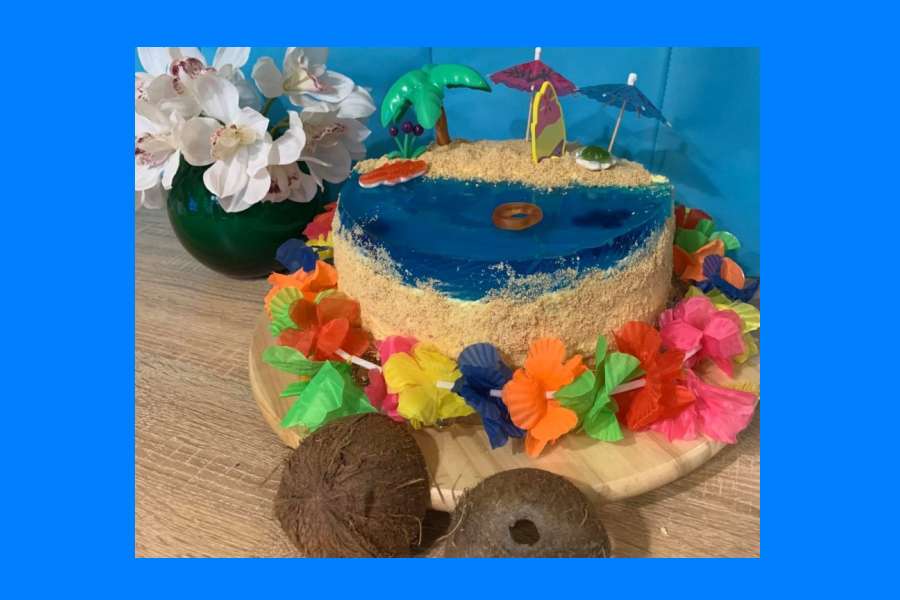 tort w kolorze niebiesko-beżowym, z hawajskimi ozdobami (kwiatami wokół) oraz palmą i parasolkami.