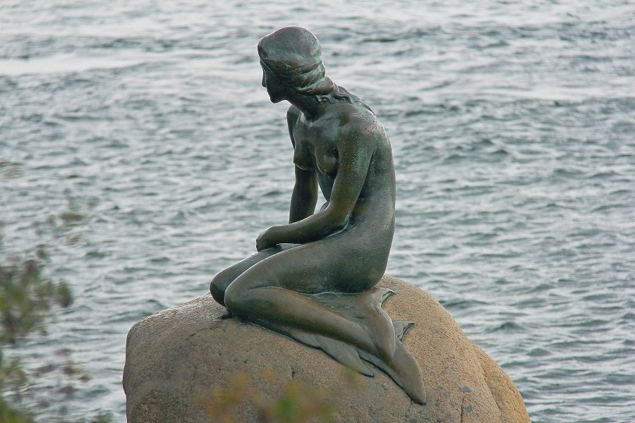 Rzeźba syreny o ludzkich nogach siedzącej na szarym głazie i wpatrującej się w stalowe morze za nią
