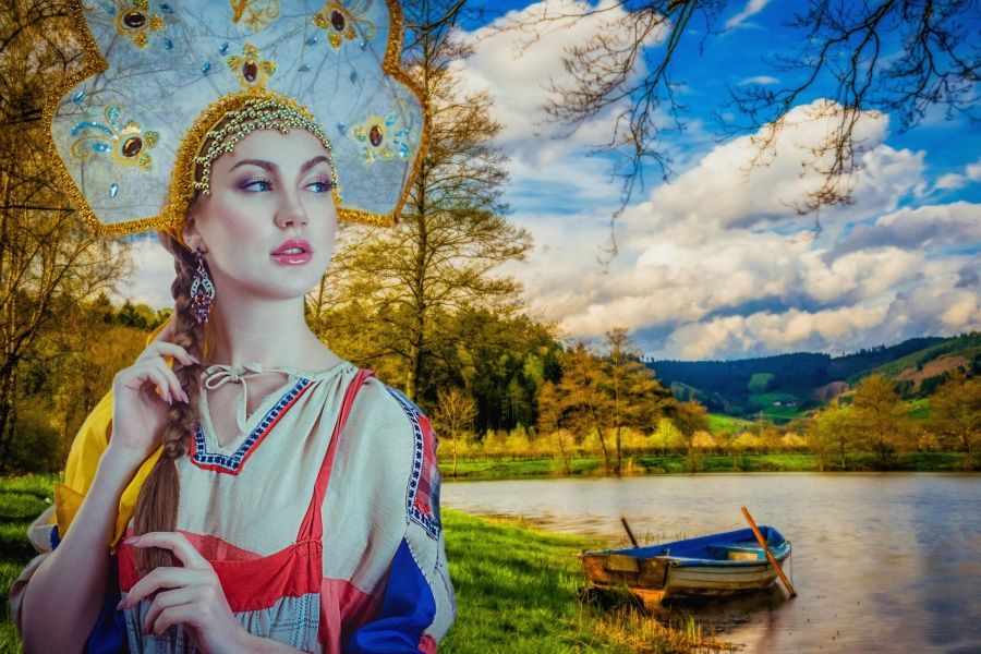 Zdjęcie przedstawia kobietę w ludowym stroju rosyjskim na tle jeziora z zacumowaną łodzią.